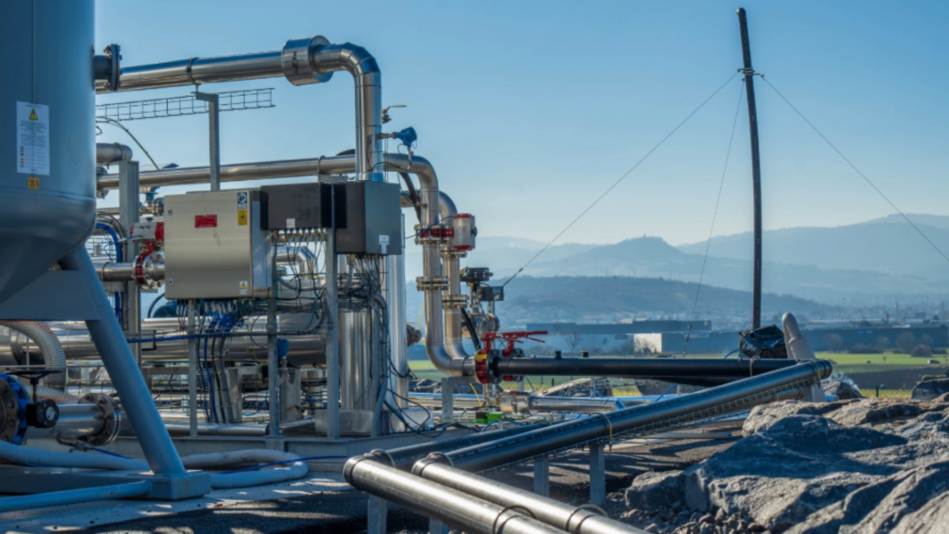 Transport du biogaz hybride Photograph of an outdoor biogas upgrading installation featuring stainless steel pipes, valves, control panels, and processing units mounted on metal structures. The facility is located in a rural landscape with hills and fields visible in the background under clear daylight, illustrating industrial equipment used to purify biogas into renewable natural gas.